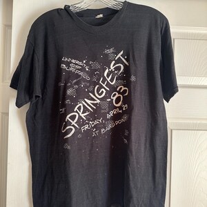May include: Black t-shirt with white text that reads "SPRINGFEST '83" and "UNIVERSITY OF BUFFALO". The shirt also includes the date "FRIDAY, APRIL 29 AT BAIRD POINT". The shirt is hanging on a clear plastic hanger.