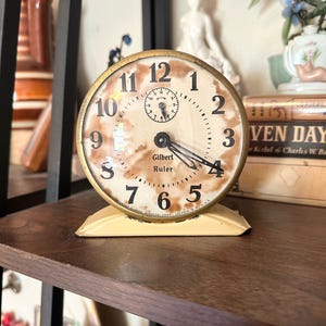 May include: An antique Gilbert Ruler clock with a cream-colored face and black numerals. The clock has a gold-colored metal frame and a small second dial. It sits on a cream-colored base. The clock is displayed on a dark wooden shelf.