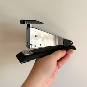 May include: A silver and black stapler. The stapler has a rectangular base and a curved top. The stapler is held in a person's hand. The stapler is made of metal and plastic.