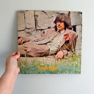 May include: Vintage vinyl record album cover featuring James Taylor. The album cover shows a man in a brown suit lying on grass in front of a stone wall. The album title and artist's name are in orange text.