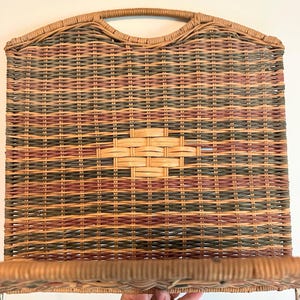 May include: Woven wicker wall shelf with a handle. The shelf features a rectangular design with a mix of brown, green, and red hues. The shelf has a woven pattern and a natural, rustic aesthetic.