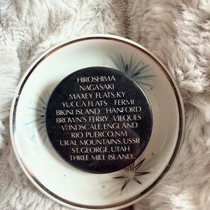 May include: A black, round object with white text listing locations like Hiroshima and Nagasaki, resting on a white plate with a gold rim and blue starburst designs. The plate sits on a fluffy, light-colored surface.