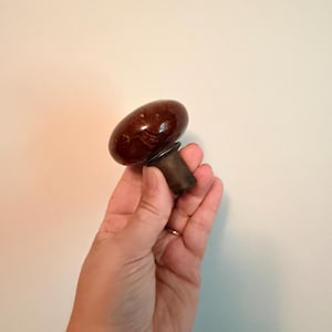 May include: A round, dark brown, polished glass or ceramic knob with a smooth, glossy finish. The knob is attached to a short, cylindrical, dark metallic base that extends outward, featuring a weathered, matte texture and a simple, functional design.