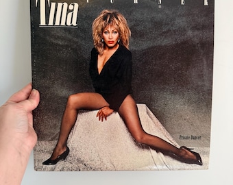 May include: A vintage vinyl record album cover featuring Tina Turner. The album cover has the name "Tina" in large white letters. Tina Turner is wearing a black outfit and fishnet stockings. The album title "Private Dancer" is visible.