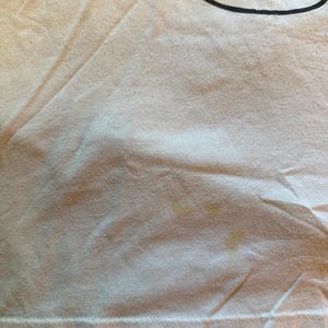 以下が含まれることがあります： 右上に黒いグラフィックデザインが施された、薄いピンク色のTシャツのクローズアップ。シャツには黄ばみがあります。生地は少し擦り切れており、質感があります。