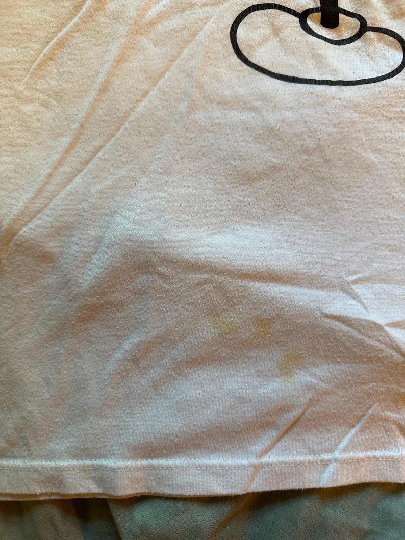 以下が含まれることがあります： 右上に黒いグラフィックデザインが施された、薄いピンク色のTシャツのクローズアップ。シャツには黄ばみがあります。生地は少し擦り切れており、質感があります。