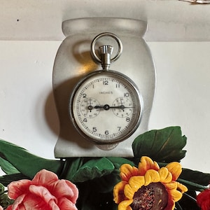 May include: A vintage pocket watch with the word "INCHES" on the face, set against a light-colored background. The watch is silver-toned with black numerals. Below the watch are colorful artificial flowers, including red roses and yellow sunflowers.