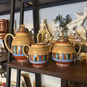 May include: A set of three decorative teapots with gold handles and lids. The teapots have a beige background with horizontal stripes in red, blue, green, and black. The teapots are arranged on a dark brown shelf.