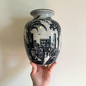 May include: A black and white ceramic vase with a detailed landscape design. The vase features a crackle glaze and a wide, rounded body. The design includes trees, buildings, and figures, creating a classic, decorative piece.