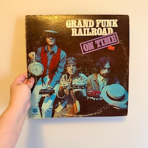 May include: Vintage vinyl record album cover for Grand Funk Railroad's "On Time." The cover features three men in retro attire, with the band's name and album title in bold text. The album cover is held by a hand.