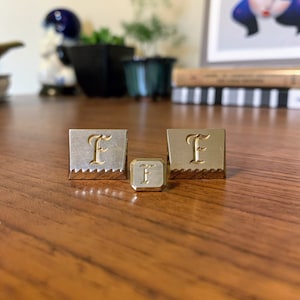 May include: Three metal objects with the letter "F" engraved on them. Two are square with a decorative edge, and one is a smaller cube. The letter "F" is in a script font and is gold-colored, contrasting with the silver and gold metal.
