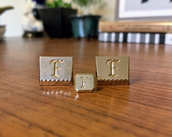 May include: Three metal objects with the letter "F" engraved on them. Two are square with a decorative edge, and one is a smaller cube. The letter "F" is in a script font and is gold-colored, contrasting with the silver and gold metal.