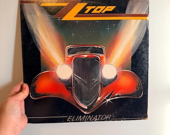 May include: Vintage album cover for ZZ Top's Eliminator. The cover features a red hot rod car with bright headlights against a starry night sky. The band's name, ZZ Top, is in bold yellow and orange letters at the top.
