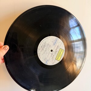 May include: A black vinyl record with a white and yellow label. The label features text and the name "Cotillion". The record is held by a hand against a light background. The record is a vintage music item.