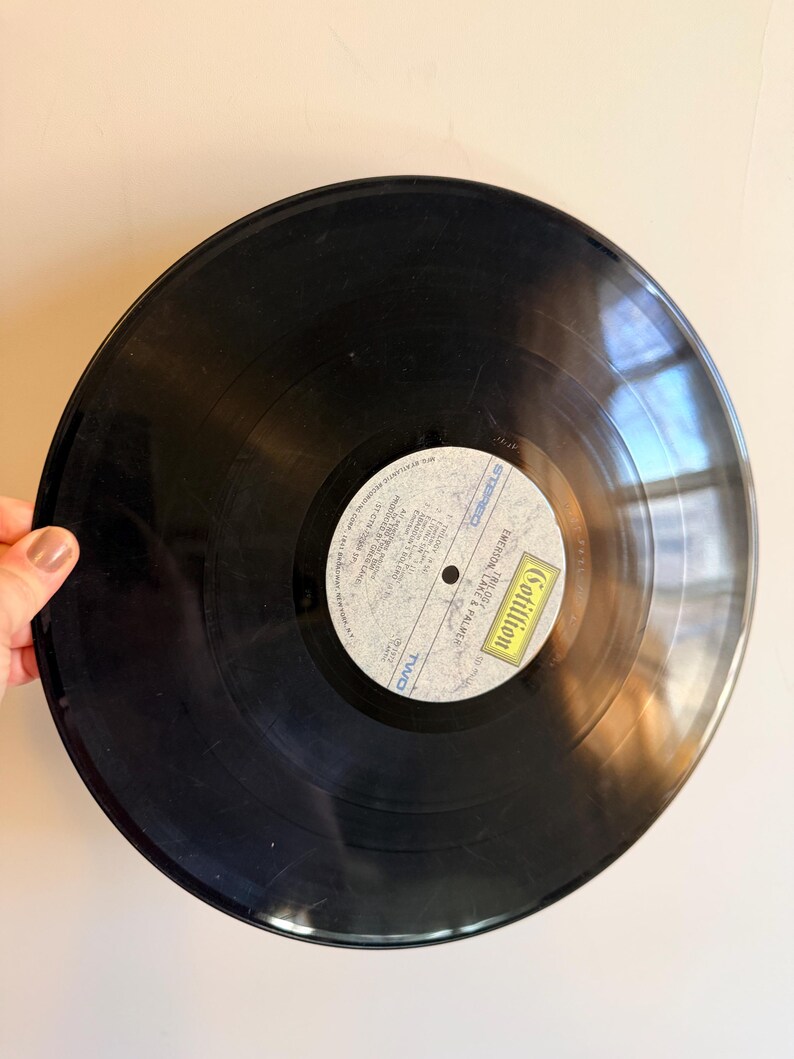 May include: A black vinyl record with a white and yellow label. The label features text and the name "Cotillion". The record is held by a hand against a light background. The record is a vintage music item.