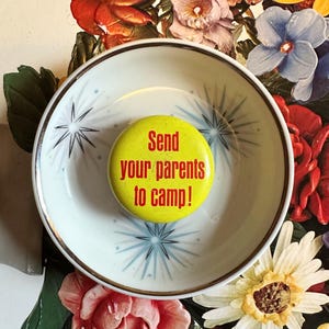 May include: A yellow button with red text that reads "Send your parents to camp!" sits in a white and gold rimmed bowl with a starburst design. The bowl rests on a floral background with red, pink, and yellow flowers.