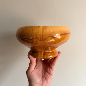 May include: A vintage, pedestal-style bowl with a glossy, amber glaze. The bowl has a wide, rounded top and a sturdy, tiered base. The glaze features darker brown drips, adding visual interest. The bowl is held by a hand.