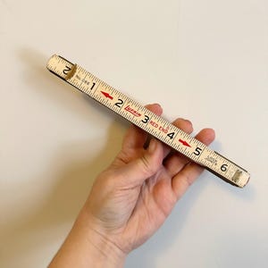 May include: A vintage Lufkin Red End folding ruler, marked with black numbers and red arrows. The ruler is white with black markings and is held in a hand. The ruler measures up to 6 inches.