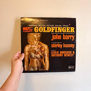 May include: Goldfinger original motion picture soundtrack vinyl record. The album cover is black with gold and orange text. The cover features a gold-painted woman and the names John Barry and Shirley Bassey.