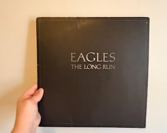May include: A square black vinyl record jacket featuring the text EAGLES THE LONG RUN in silver lettering centered on the front cover. The album cover has a matte finish with a thin embossed border line running near the edges of the sleeve.