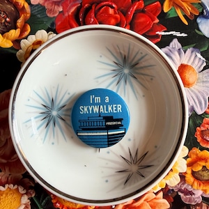 May include: A white ceramic bowl with a gold rim and blue starburst designs. A blue button with the text "I'm a SKYWALKER" and "PRUDENTIAL" rests inside the bowl. The background features a floral pattern with red, orange, and yellow flowers.