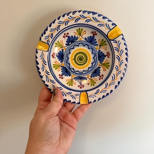 May include: Hand-painted ceramic ashtray with a floral design. The ashtray features a white base with blue, yellow, and green accents. The center has a spiral design, and the rim has three yellow rests.