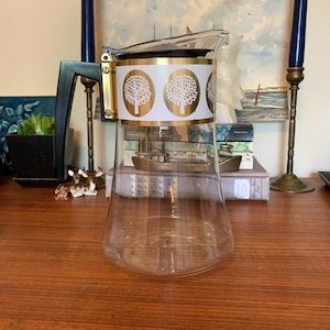 May include: A clear glass coffee pot with a gold and white band featuring a repeating pattern of stylized trees. The pot has a black plastic handle.