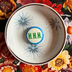 May include: A white ceramic bowl with a silver rim and a blue and black starburst pattern. Inside the bowl is a round button with the letters "H.H.H." in green on a white background, surrounded by a blue border. The background is a floral pattern.