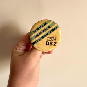 May include: A round, yellow IBM DB2 button with text in black and blue. The text includes phrases like "the world is flat" and "man will never fly." The button has a vintage, slightly worn appearance.