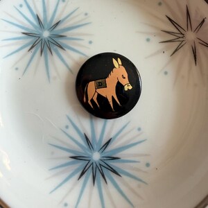 May include: A black button with a cartoon donkey in orange and yellow, with the letter "D" on its side. The button is set in a white bowl with a blue starburst pattern and a brown rim.