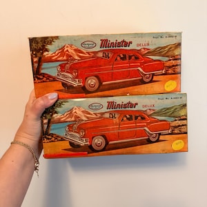May include: Two rectangular cardboard boxes for a Minister Delux toy car. Each box features a vintage illustration of a red sedan against a mountain landscape, with the text Minister Delux, Mechanical Automatic, and Regd. No. A-34951-81 printed on them.