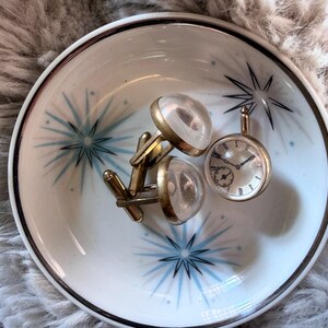 May include: A white and blue decorative dish with starburst designs holds a pair of gold cufflinks with clear, round tops and a small, round pocket watch. The dish has a dark brown rim.