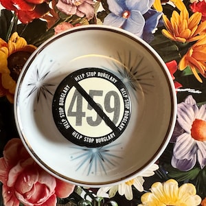 May include: A white bowl with a gold rim and starburst design holds a black and white circular object. The object has the text "HELP STOP BURGLARY" and the number "459" crossed out. The background is a floral pattern.