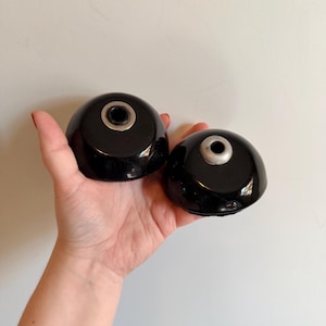May include: Two black, dome-shaped objects with a silver metal ring on top. The objects are held in a hand against a white background. The objects appear to be decorative or functional, possibly for a home decor or kitchen item.