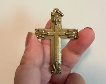 May include: A gold-colored cross pendant with decorative leaf accents on each arm. The cross is held up by a hand against a white background. The pendant has a small loop at the top for a chain.