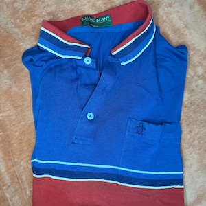 May include: Blue polo shirt with red collar and hem. Features a white and blue striped collar and a small pocket with a penguin logo. The shirt has two buttons and the tag reads "GRAND SLAM".
