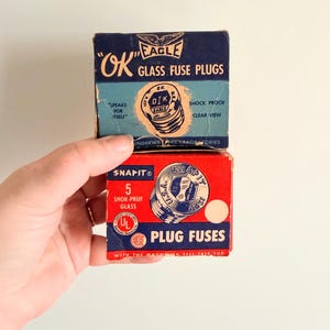 May include: Two vintage boxes of glass fuse plugs. The top box is navy blue with the text "OK EAGLE GLASS FUSE PLUGS". The bottom box is red with the text "SNAPIT PLUG FUSES". Both boxes are held by a hand.
