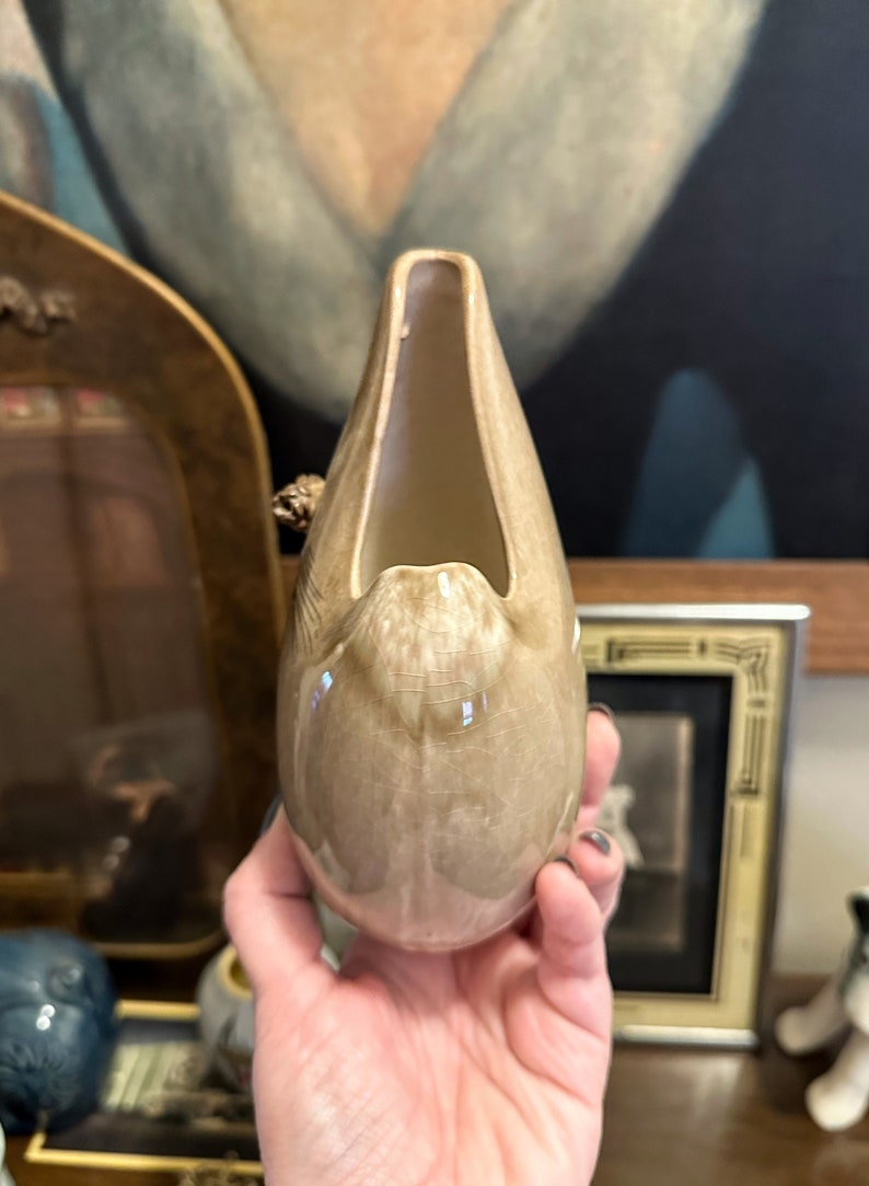May include: A light brown ceramic vase shaped like a bird's head with a wide open beak. The vase has a speckled glaze and a glossy finish.