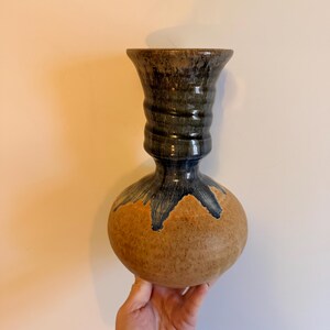 May include: A handcrafted ceramic vase with a bulbous base and a flared, textured neck. The vase features a speckled tan body with dark blue and brown accents, creating a unique, artistic aesthetic. The vase is held by a hand.