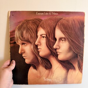 May include: Vintage vinyl record album cover for Emerson, Lake & Palmer's "Trilogy." The cover art features three side profiles of individuals with long hair. The album title and band name are printed on the cover.