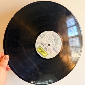 May include: A black vinyl record with a white and yellow label. The label features text including "Cotillion" and "Emerson, Lake & Palmer". The record is held by a hand.