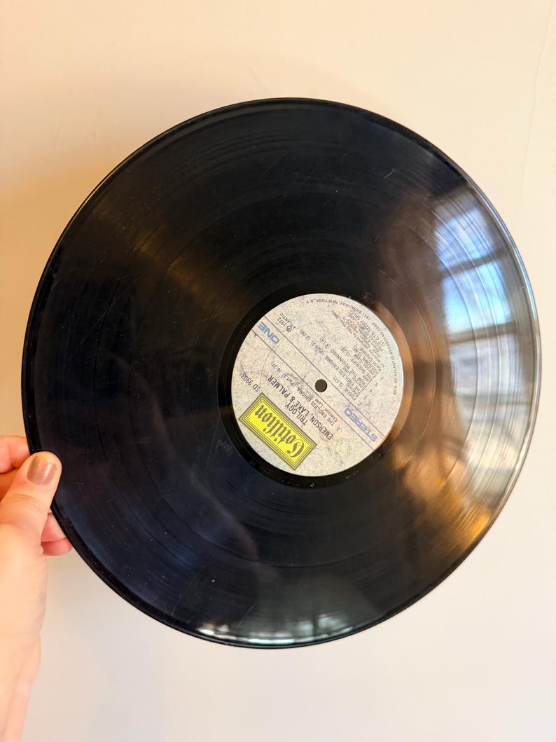 May include: A black vinyl record with a white and yellow label. The label features text including "Cotillion" and "Emerson, Lake & Palmer". The record is held by a hand.