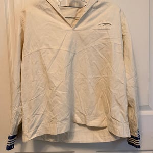 May include: A long-sleeved off-white pullover shirt featuring a sailor-style collar, a V-neckline, and horizontal navy blue and white striped cuffs. The garment has a horizontal seam across the chest and is displayed on a clear plastic hanger.