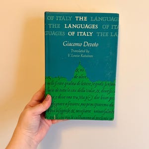 May include: A teal-colored book titled "The Languages of Italy" by Giacomo Devoto. The book's cover features text in white and green, with a green outline of Italy. The book is held by a person.
