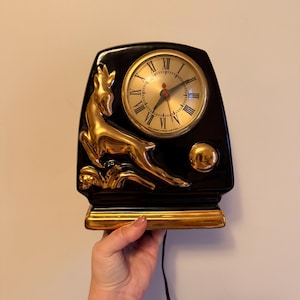 May include: Black and gold ceramic clock with a deer motif. The clock face is gold with black Roman numerals. The deer is gold, and the clock has a gold base and accents. The clock is being held by a person.