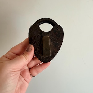 May include: A vintage, heart-shaped padlock with a dark, weathered finish. The lock features a curved handle and a rectangular keyhole cover. The metal has a rusty, aged appearance, suggesting it is an antique.