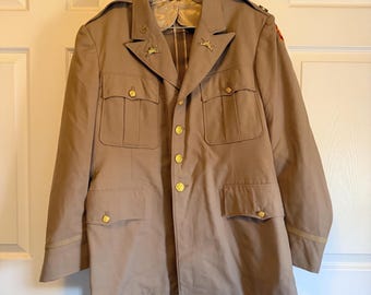 May include: A tan military jacket with gold buttons and pockets. The jacket has a notched collar and epaulettes. Two gold star pins are on the lapel. The jacket is hanging on a hanger against a white door.