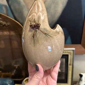 May include: A light brown ceramic vase with a unique teardrop shape and a pine cone design. The vase has a crackled glaze and a small opening at the top.