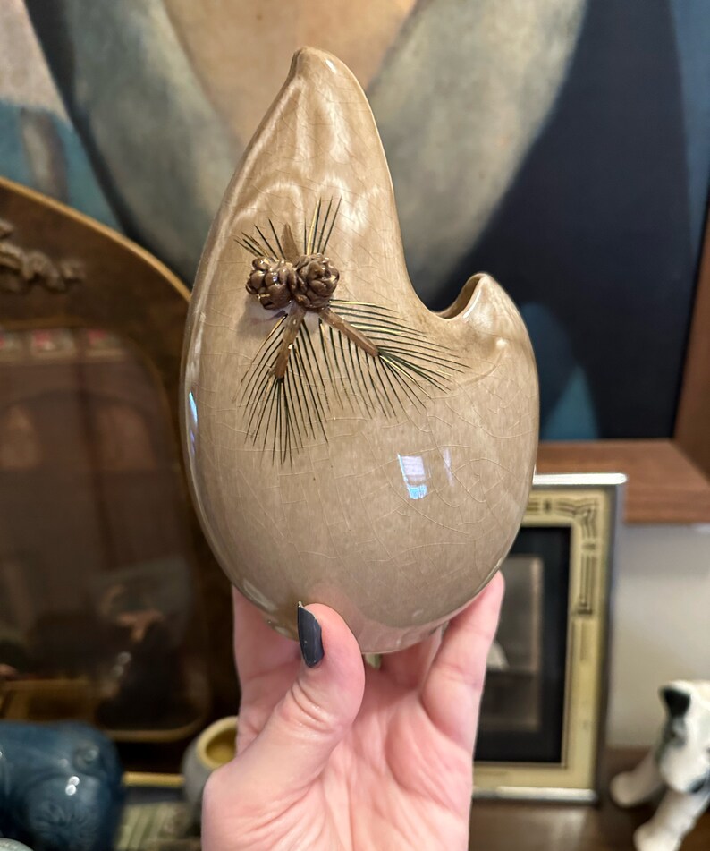 May include: A light brown ceramic vase with a unique teardrop shape and a pine cone design. The vase has a crackled glaze and a small opening at the top.