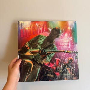 May include: A vinyl record album titled "Buzzcocks: A Different Compilation." The album cover features a band performing live, with the band name in large red letters. The image is wrapped in clear plastic.